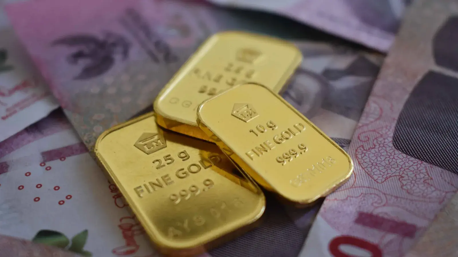 Gold bullion bars stamped with refinery hallmarks on a stainless steel assay table, with a technician recording weights alongside