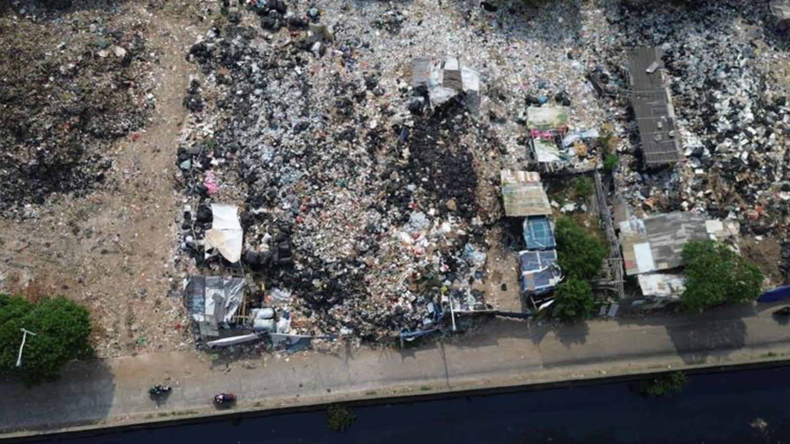 City sanitation trucks and heavy equipment clearing a long stretch of illegally dumped waste along a green open space in North Jakarta
