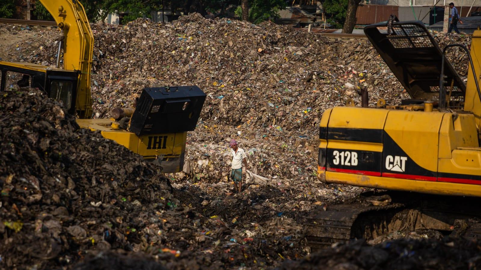 Truk sampah mengantre di area kerja TPA tempat sampah dipadatkan dan ditutup tanah, dengan ekskavator dan material penutup terlihat di latar belakang