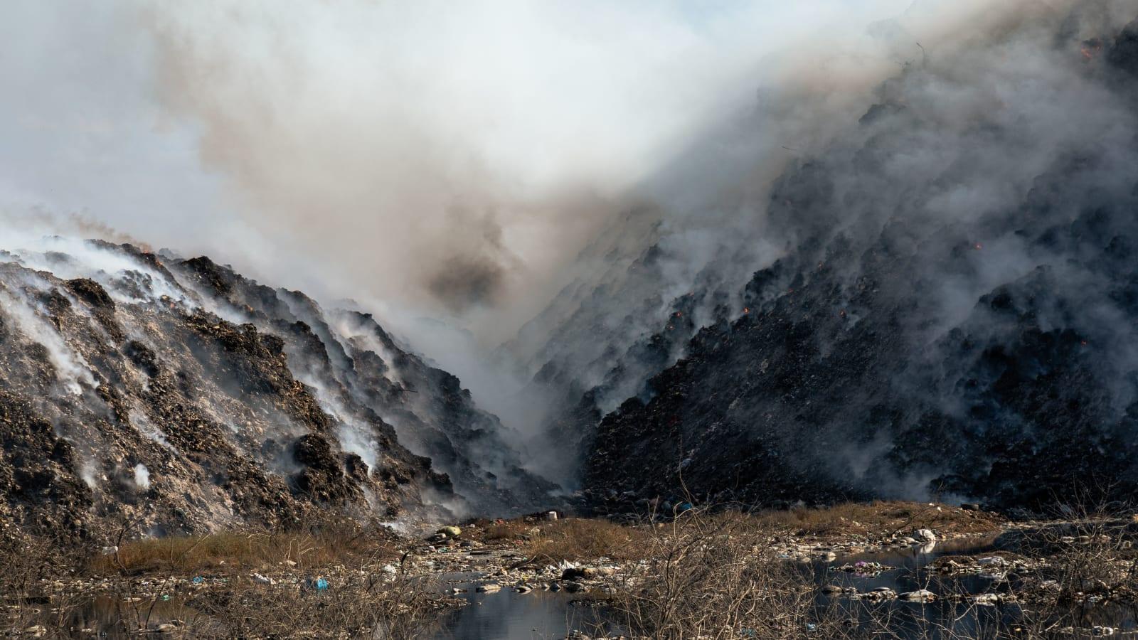 Firefighters and water tanker trucks working at night to contain a fire at the edge of a large municipal landfill, with an excavator standing by