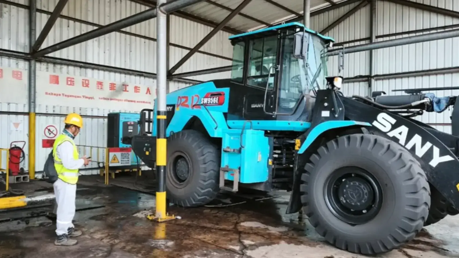 Ekskavator listrik memuat bijih ke dump truck listrik di area stockpile di samping bangunan smelter, dengan stasiun pengisian daya di latar depan